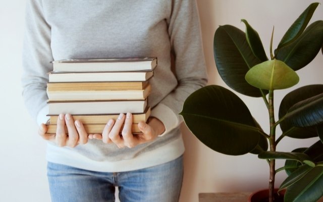 a girl standing by a wall and holding a few books with both of her hands